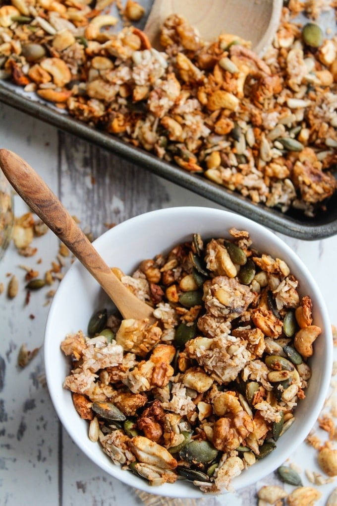 homemade banana granola made with mixed nuts, seeds and coconut in a cereal bowl with a baking sheet full of granola in the back