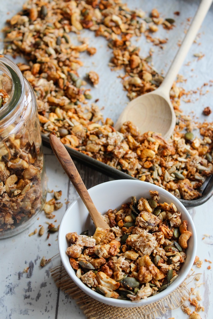 homemade banana granola made with mixed nuts, seeds and coconut in a cereal bowl with a baking sheet and storage jar full of granola in the back 