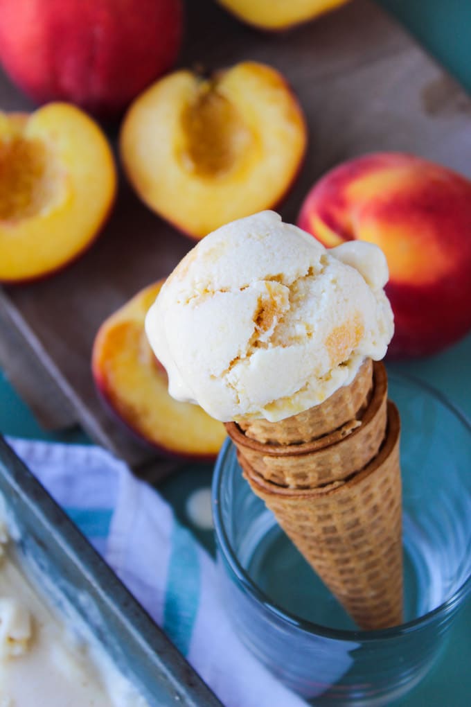 a waffle cone with a scoop of homemade peach coconut milk ice cream sitting in a cup with fresh peaches in the back