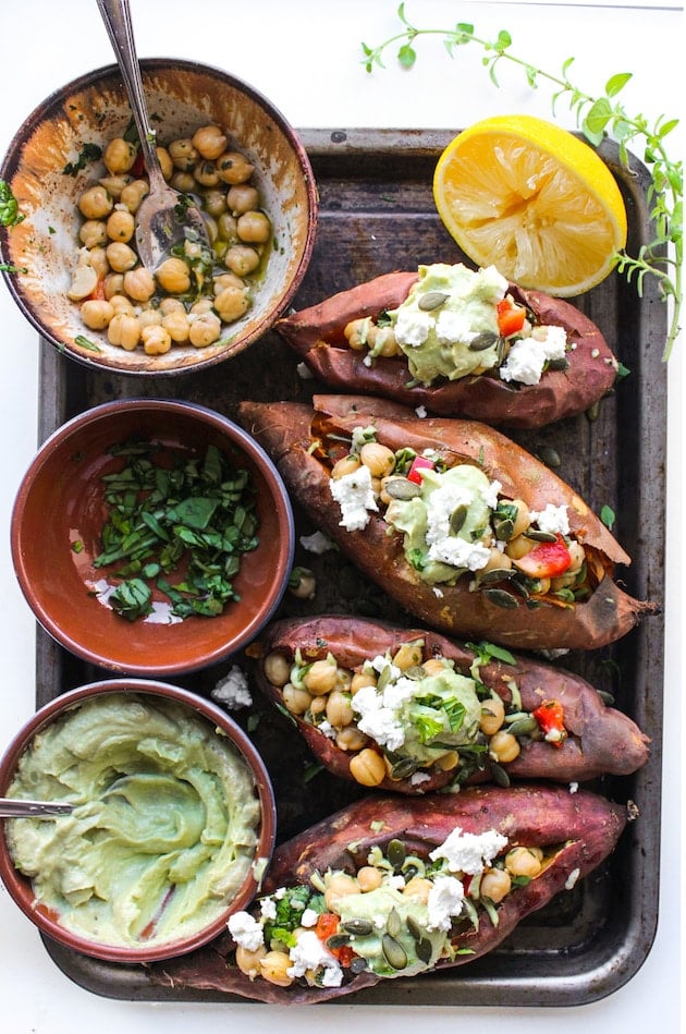 Mediterranean stuffed sweet potatoes on a tray with herbs, avocado tahini and chickpeas