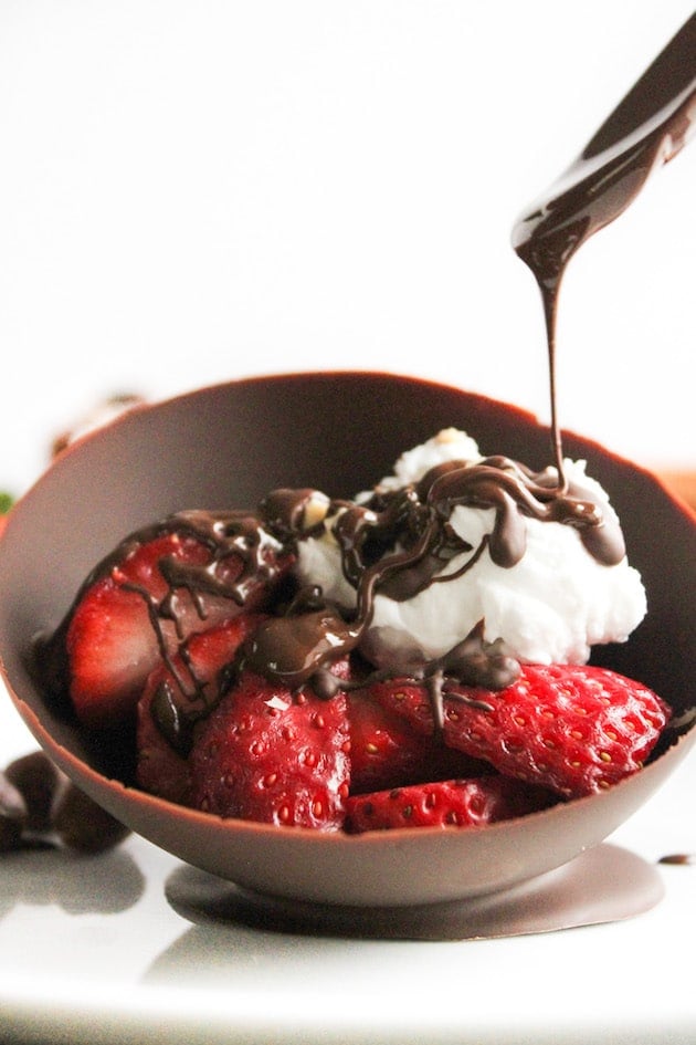 Homemade edible chocolate bowls filled with strawberries and coconut whipped cream with chocolate pouring over the top