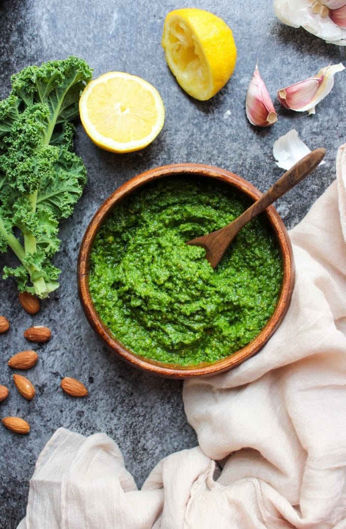 kale pesto in a bowl surrounded by the fresh ingredients (lemon, garlic, almonds and kale)