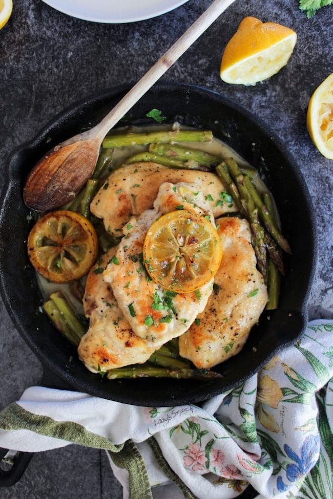 One Pan Lemon Chicken in a cast iron skillet with asparagus topped with grilled lemons and parsley and a wooden spoon 