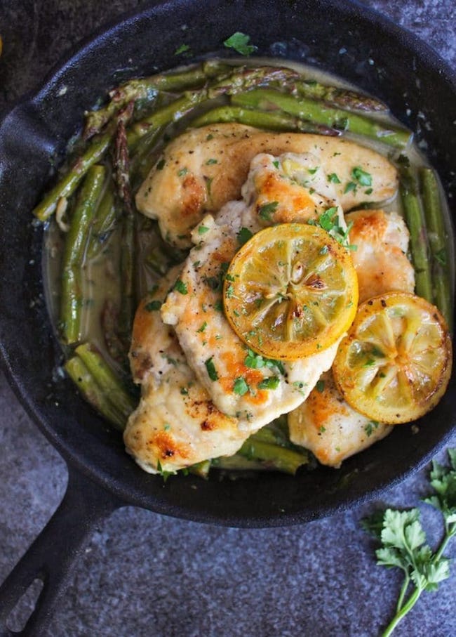 One Pan Lemon Chicken in a cast iron skillet topped with grilled lemons and parsley with asparagus