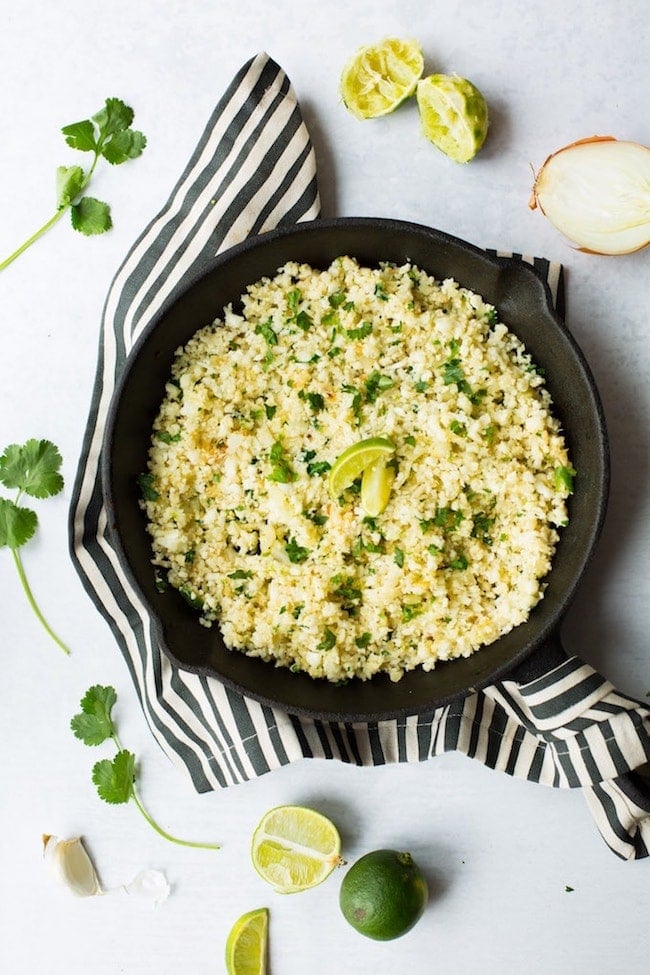 Cilantro Lime Cauliflower Rice in a cast iron skillet surrounded by limes and cilantro 