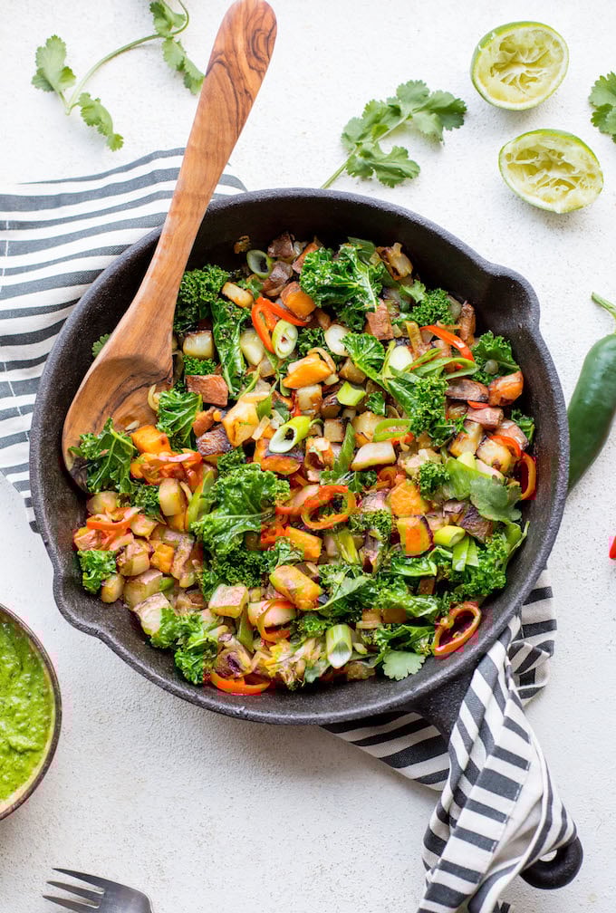 a skillet full of Kale Potato Breakfast Hash surrounded by fresh ingredients: limes, cilantro and chili peppers