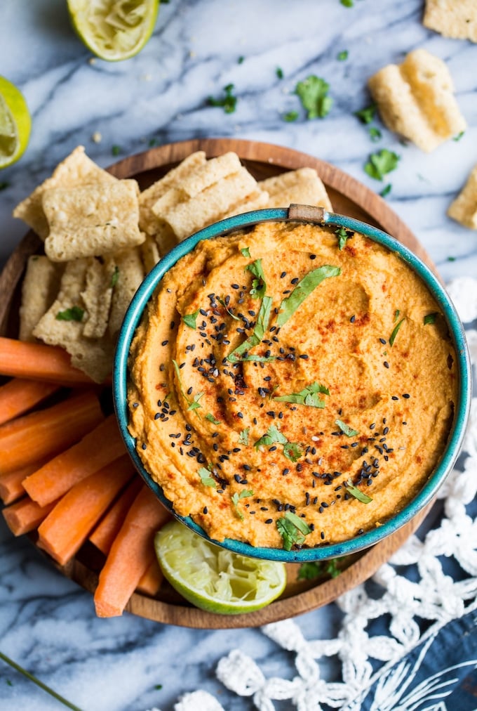 Sriracha Lime Hummus topped with black sesame seeds, chopped cilantro and paprika on a plate with hummus chips and slices carrot sticks