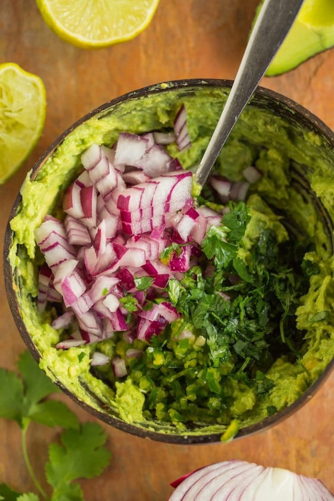 smashed avocado in a bowl with diced red onion and cilantro to make homemade guacamole