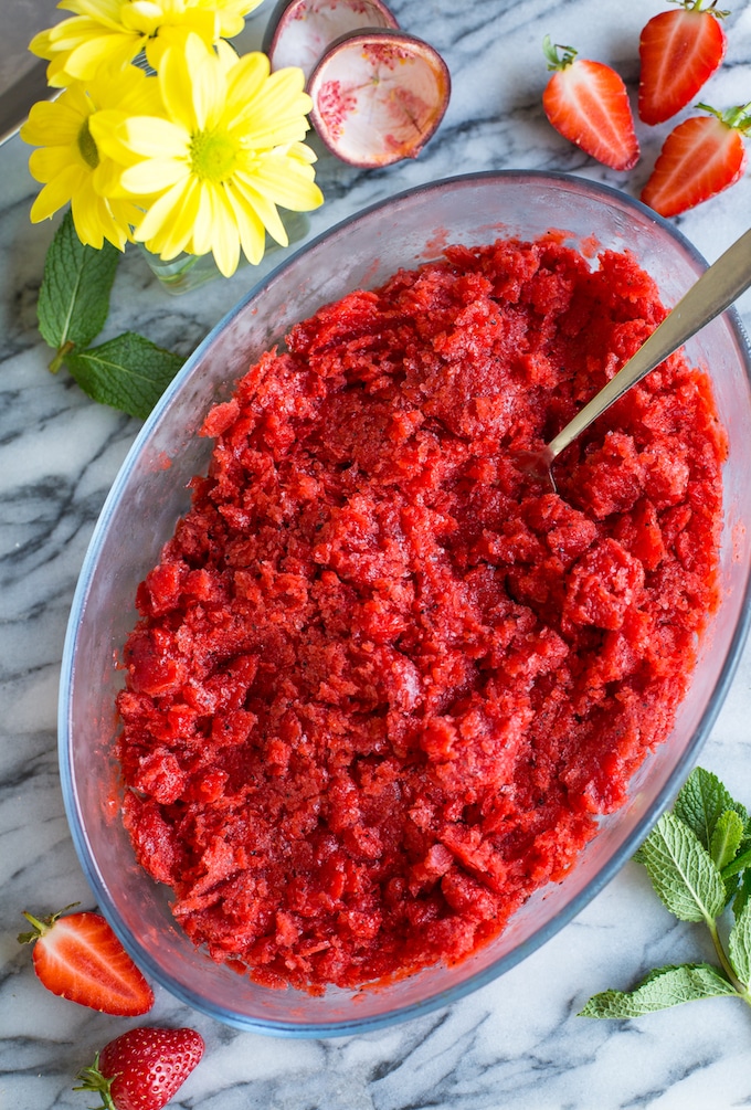 Strawberry Granita with Passion Fruit in a glass container surrounded by sliced berries and flowers 