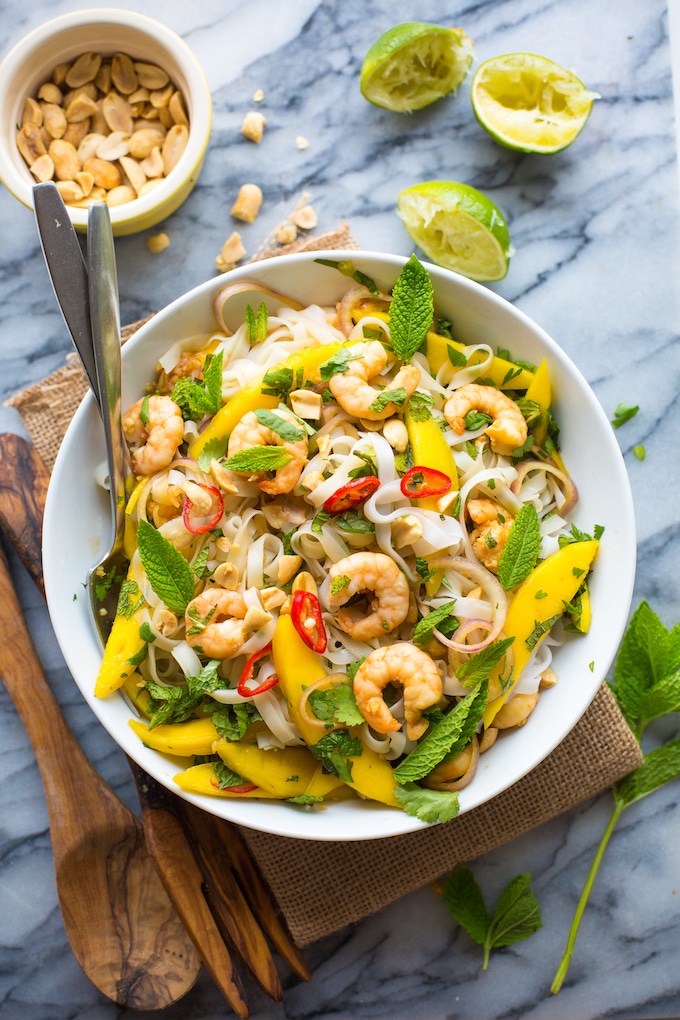 Spicy Mango & Shrimp Thai Noodle Salad in a bowl topped with fresh mint & chopped peanuts surrounded by limes
