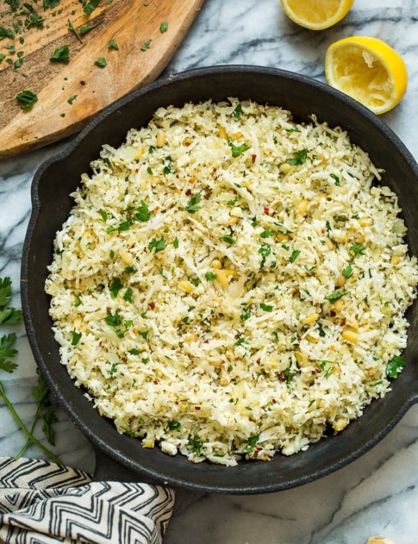 Mediterranean Cauliflower Rice in a cast iron skillet on a marble surface with lemon and parsley surrounding