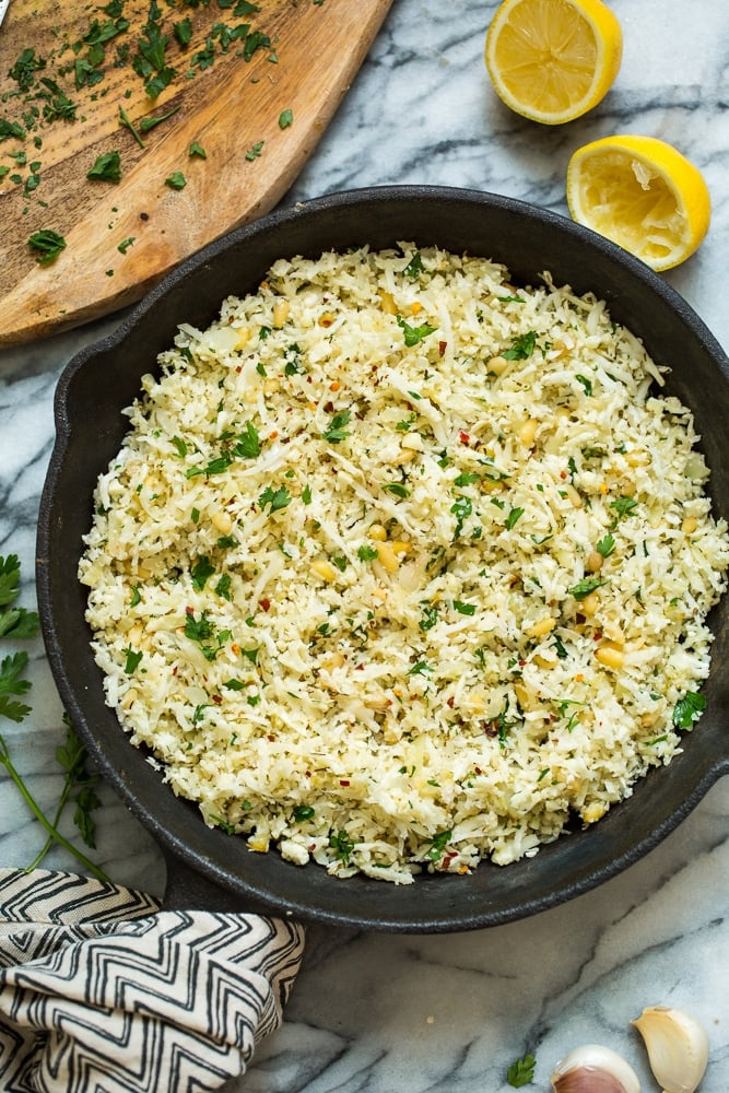  Mediterranean Cauliflower Rice in a cast iron skillet on a marble surface with lemon and parsley surrounding