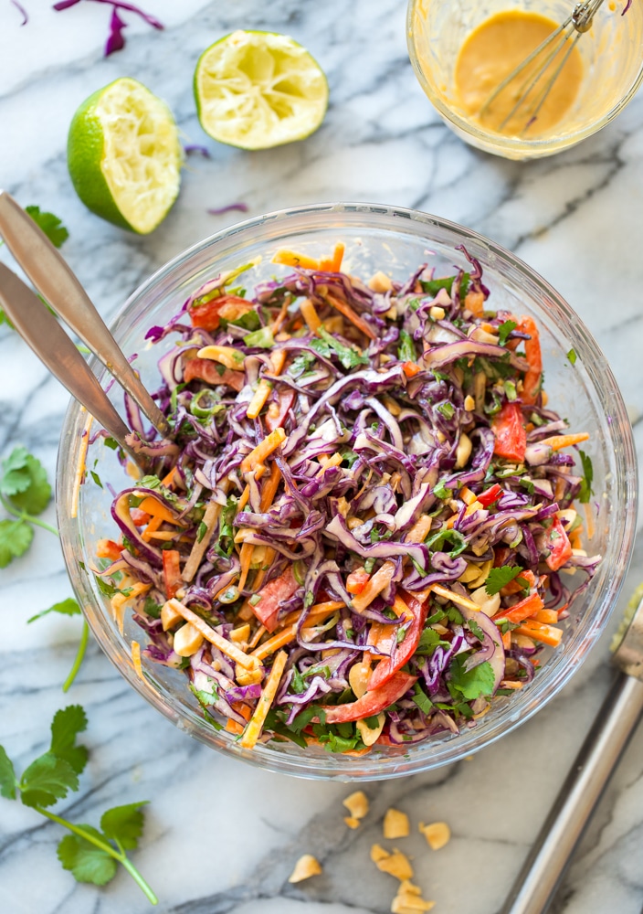 Thai Peanut Coleslaw in a salad bowl on a marble counter top surrounded by limes, fresh cilantro and chopped peanuts