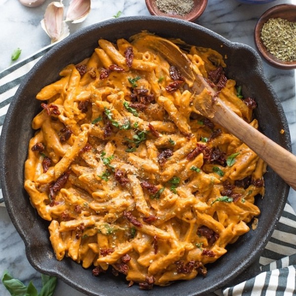 Vegan Sun Dried Tomato Alfredo in a cast iron pan surrounded by garlic cloves, oregano, fresh basil and sun dried tomatoes