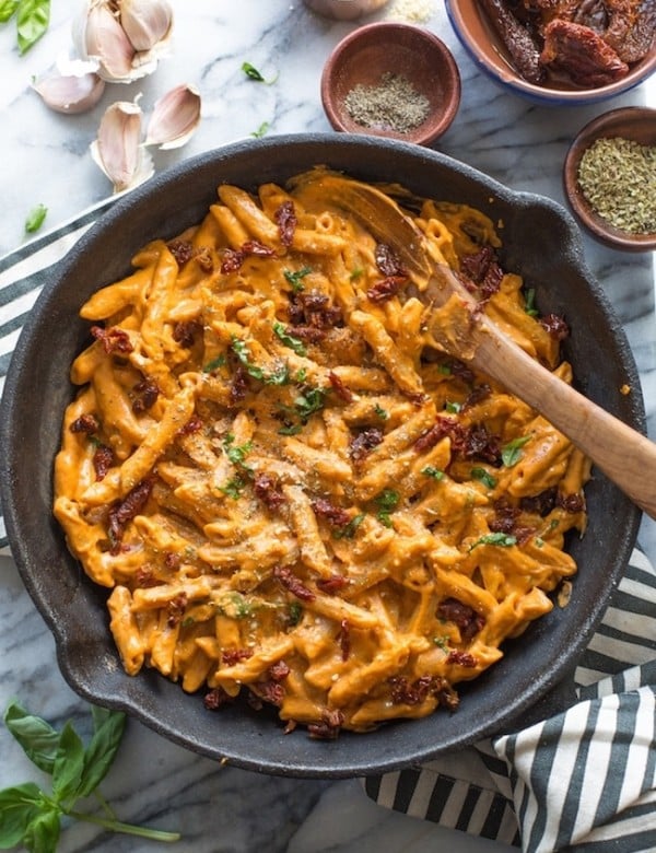 Vegan Sun Dried Tomato Alfredo in a cast iron pan surrounded by garlic cloves, oregano, fresh basil and sun dried tomatoes