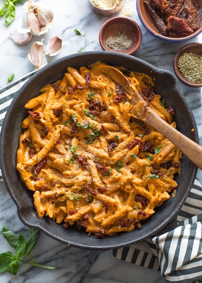 Vegan Sun Dried Tomato Alfredo in a cast iron pan surrounded by garlic cloves, oregano, fresh basil and sun dried tomatoes