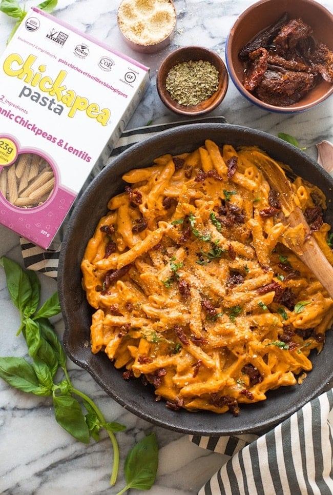 Vegan Sun Dried Tomato Alfredo in a cast iron skillet on top of a black and white stripped kitchen towel with fresh basil and chickapea pasta