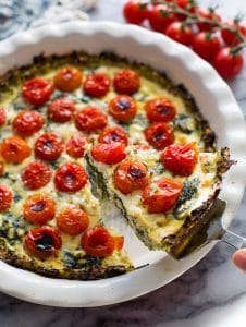Zucchini Crust Tomato Spinach Feta Quiche in a pie plate with a slice lifted up on a spatula