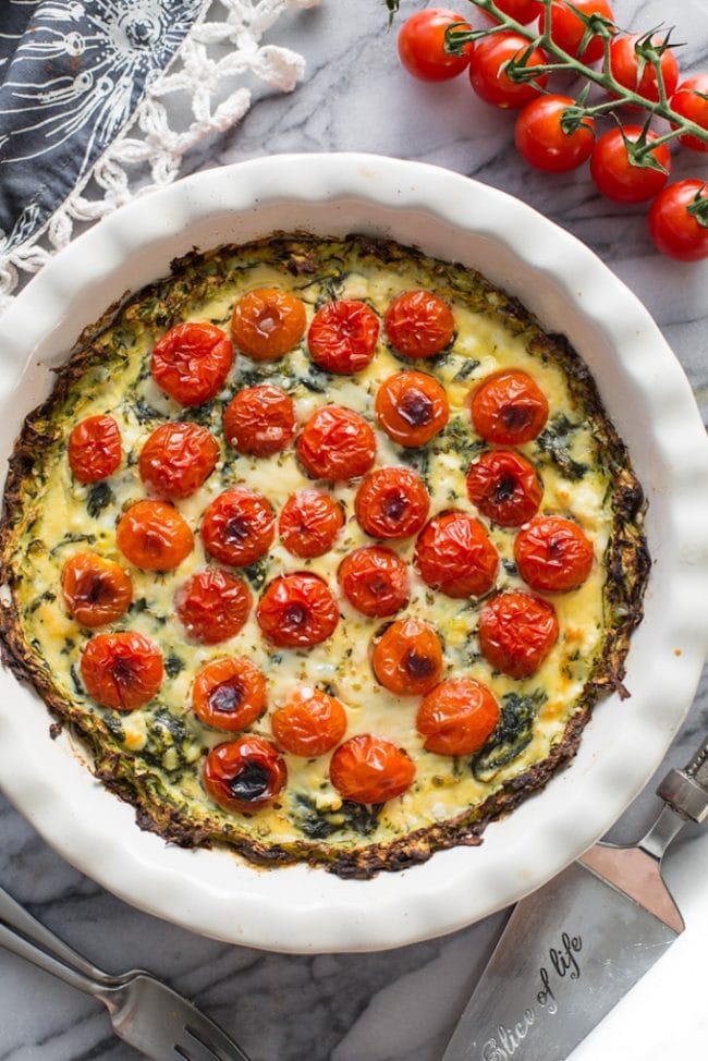 a freshly baked spinach feta pie topped with blistered cherry tomatoes over a zucchini crust in a pie dish