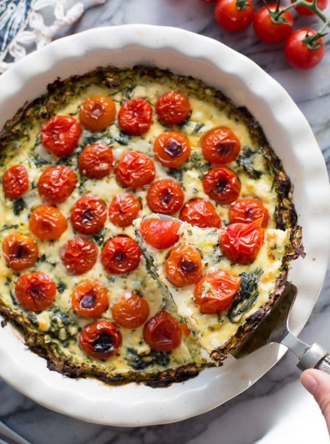 Zucchini Crust Tomato Spinach Feta Quiche in a pie plate with a spatula holding a slice