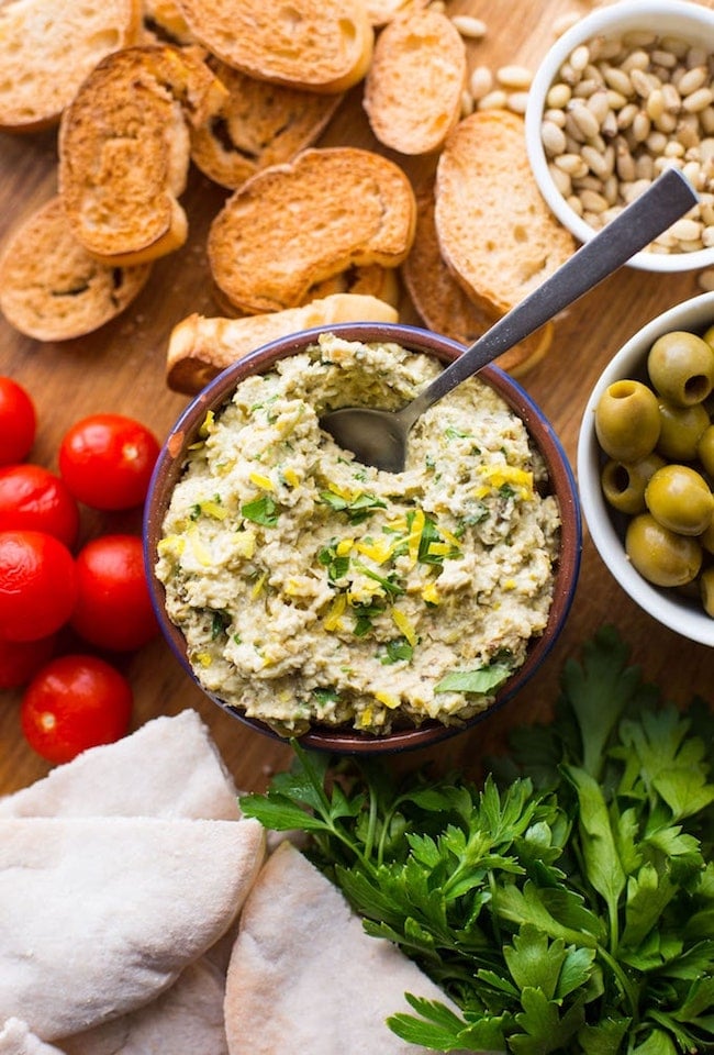 green olive tapenade on a serving board surrounded by veggies, crostini and pita