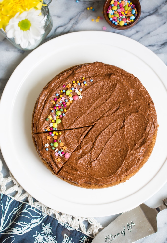 Paleo cake made with coconut flour, frosted with chocolate frosting and topped with sprinkles on a white cake plate surrounded by more sprinkles, a serving knife and flowers