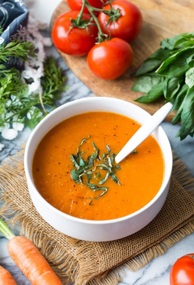 Low FODMAP Carrot Tomato Soup in a white bowl topped with fresh basil and surrounded by produce