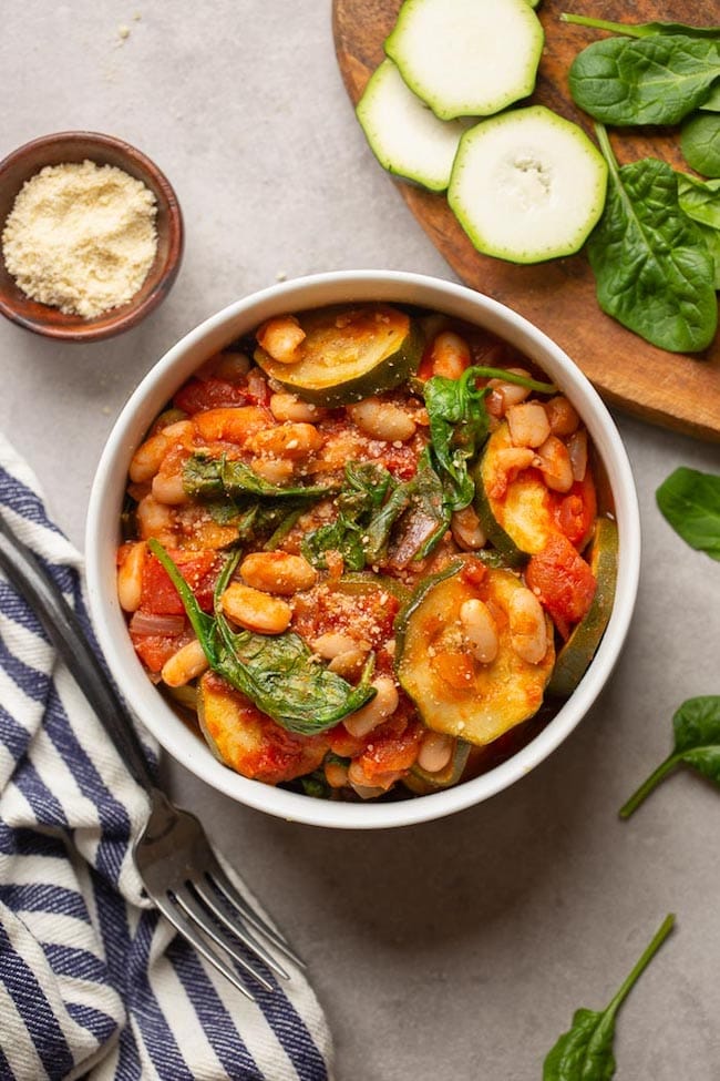 Zucchini and Cannellini Tomato Stew in a white bowl surrounded by spinach and zucchini