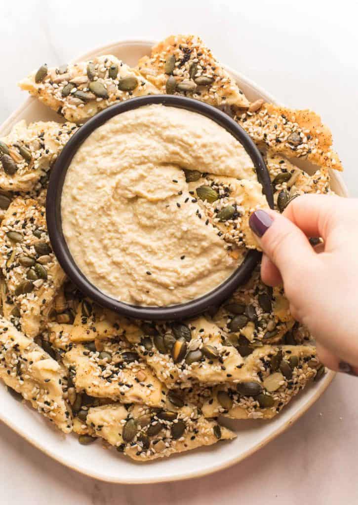 Seedy Everything Seasoned Crackers on a plate with a cracker dipping into hummus