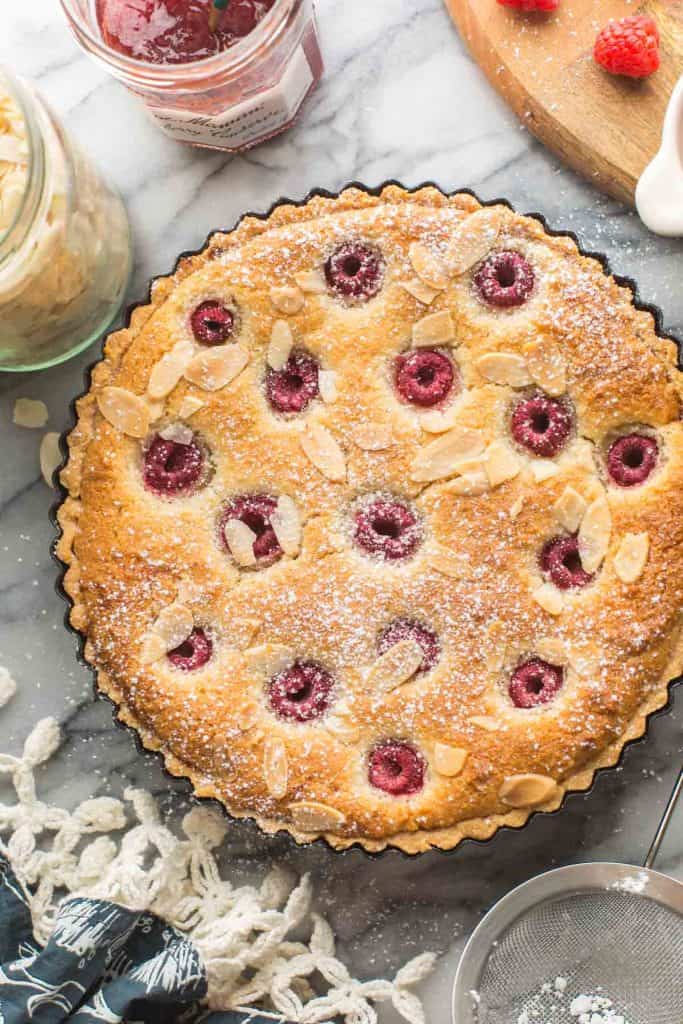 vegan bakewell tart still in the tart pan surrounded by jam, almond flakes and kitchen towel