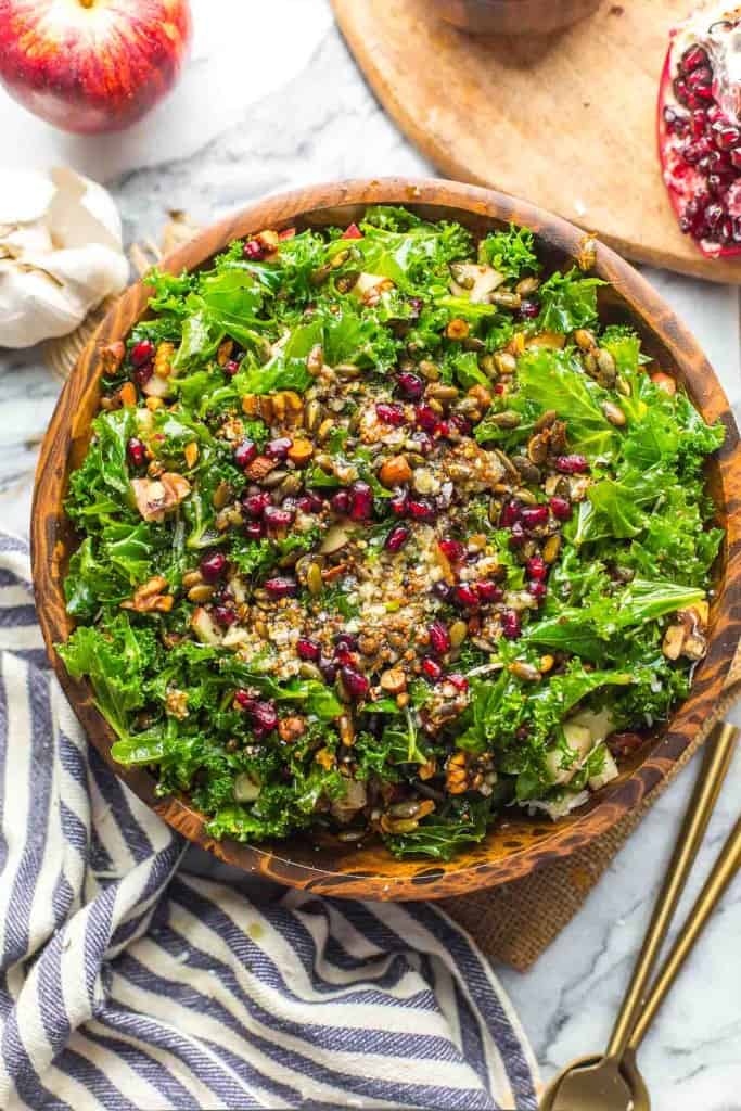 Winter Kale Salad in a wood bowl with serving utensils and a kitchen towel on the side