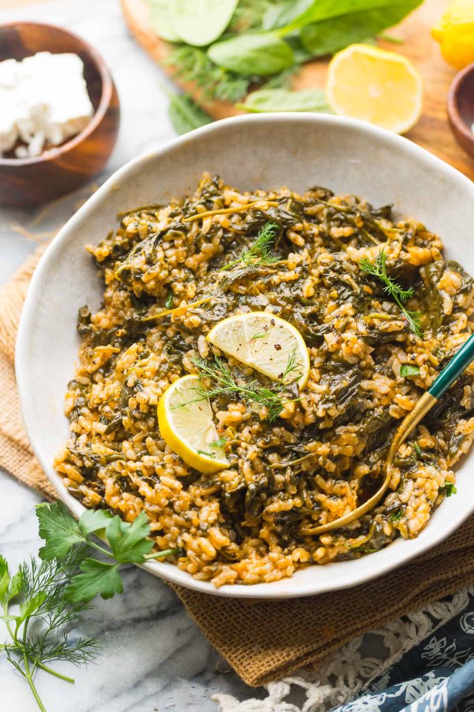 Spanakorizo in a serving bowl topped with lemon slices and fresh herbs and serving spoons