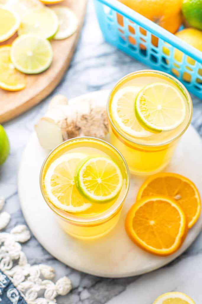 a couple of glasses of Homemade Electrolyte Drink garnished with lemon, lime and orange slices in front of a basket of citrus fruit and a cutting board with fruit slices