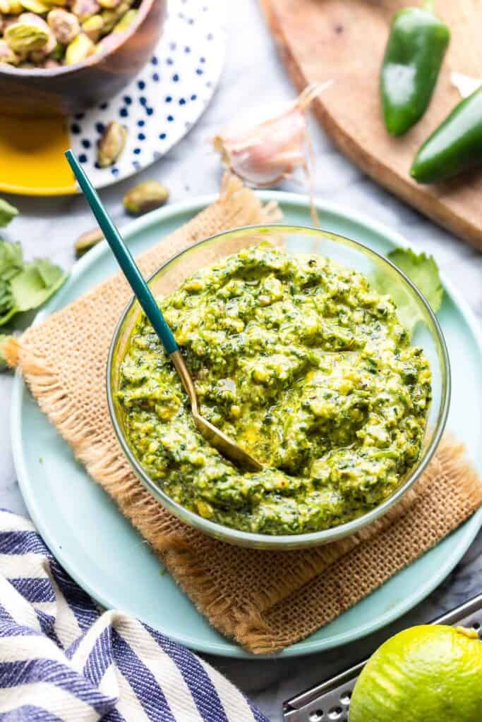 Cilantro Lime Pesto in a bowl with a spoon on a plate with a small bowl of pistachios and a couple of jalapenos behind