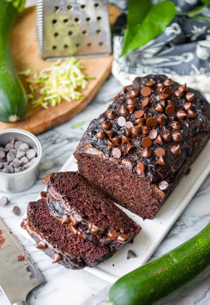 gluten free chocolate zucchini bread loaf sliced on a serving board surrounded by whole zucchini and chocolate chips