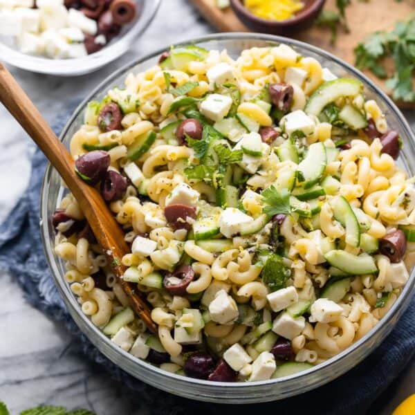 Greek Lemon & Cucumber Pasta Salad in a large salad bowl with a serving spoon surrounded by the fresh ingredients