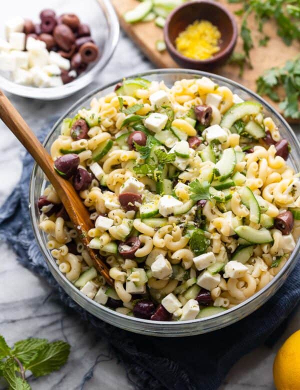 Greek Lemon & Cucumber Pasta Salad in a large salad bowl with a serving spoon surrounded by the fresh ingredients