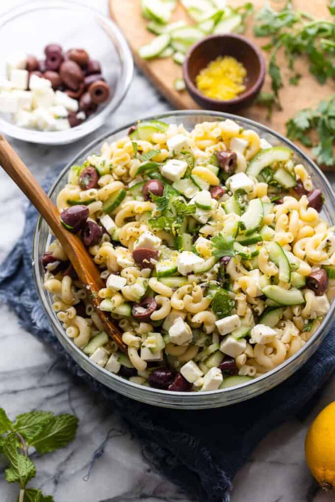 Greek Lemon & Cucumber Pasta Salad in a large salad bowl with a serving spoon surrounded by the fresh ingredients