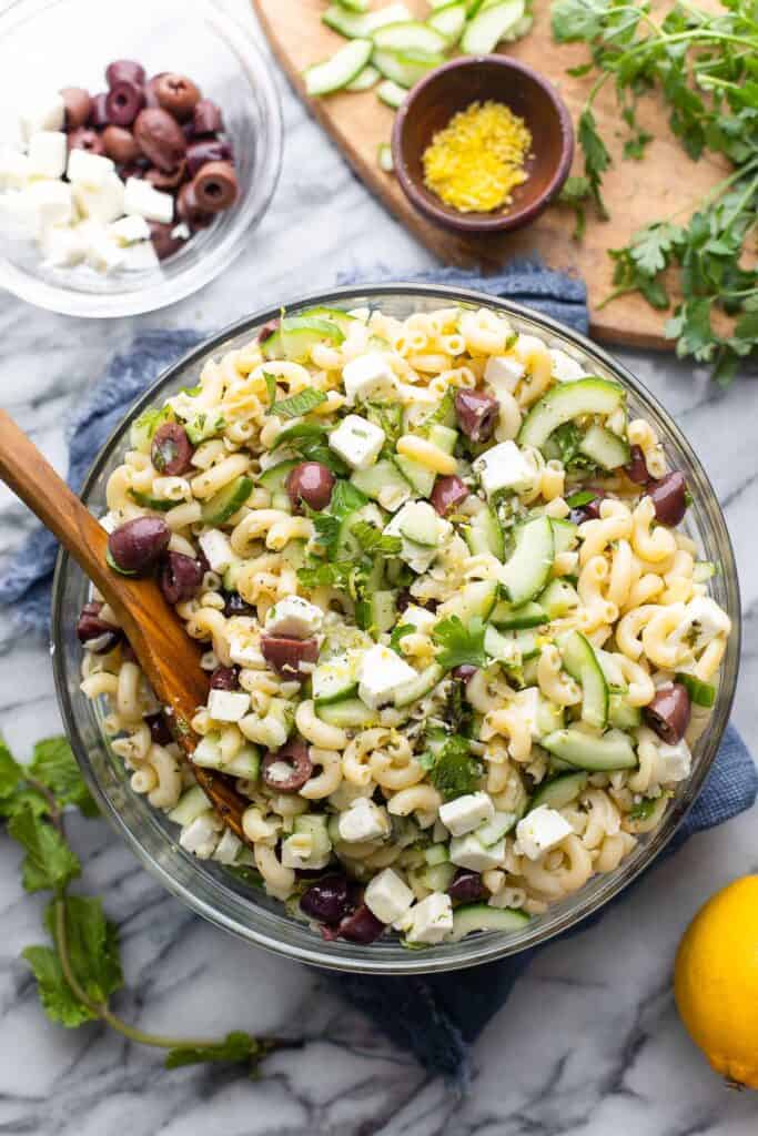 Greek Lemon & Cucumber Pasta Salad in a large salad bowl with a serving spoon surrounded by the fresh ingredients