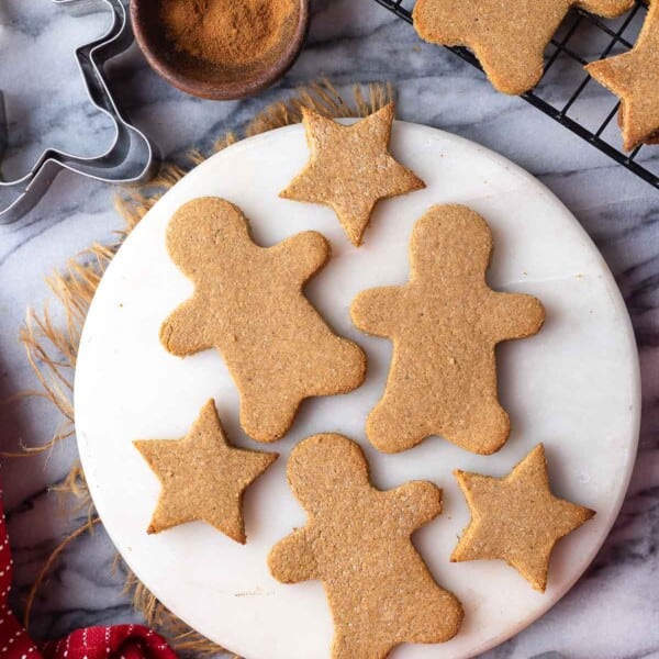 paleo gingerbread cookies in the shape of gingerbread men and stars on a round platter with cookies cooling on a wire rack in the back