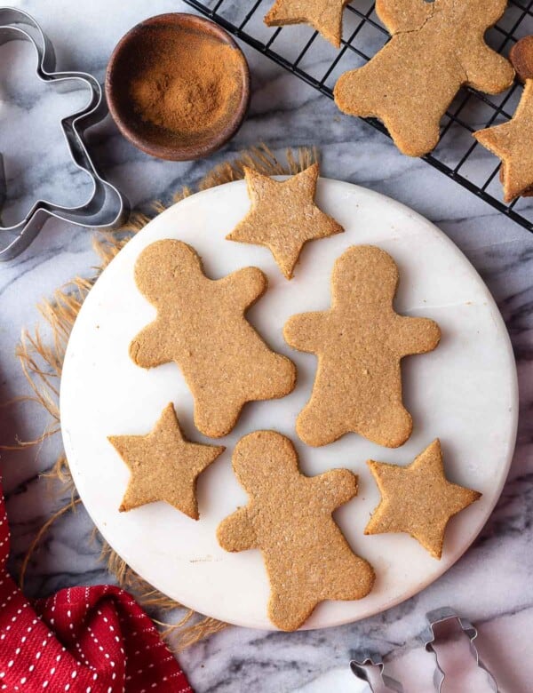 paleo gingerbread cookies in the shape of gingerbread men and stars on a round platter with cookies cooling on a wire rack in the back