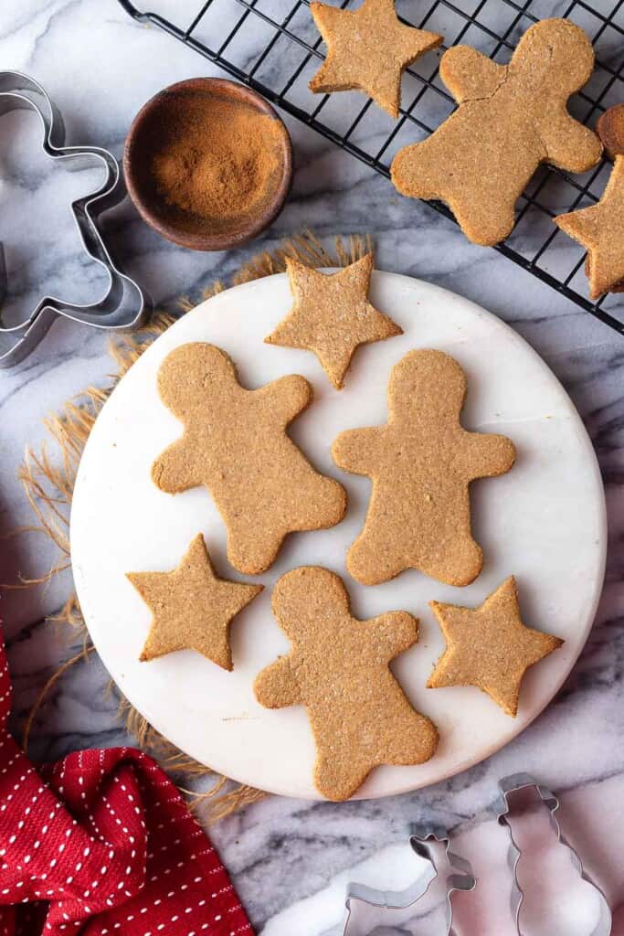 paleo gingerbread cookies in the shape of gingerbread men and stars on a round platter with cookies cooling on a wire rack in the back