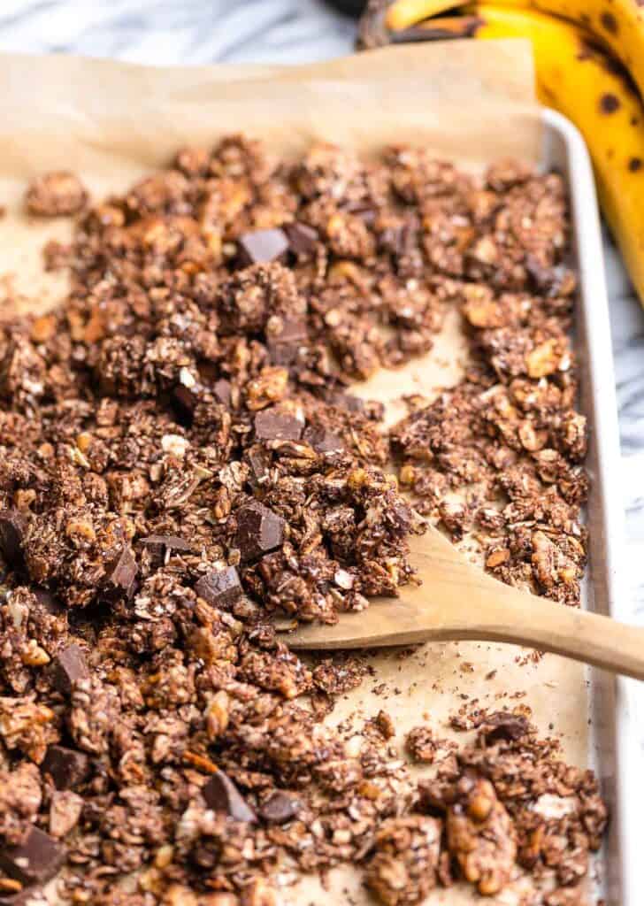 a wooden spoon scooping up homemade Banana Chocolate Granola on a baking sheet 