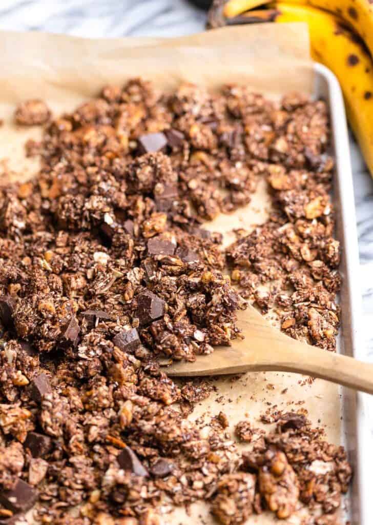 a wooden spoon scooping up homemade Banana Chocolate Granola on a baking sheet