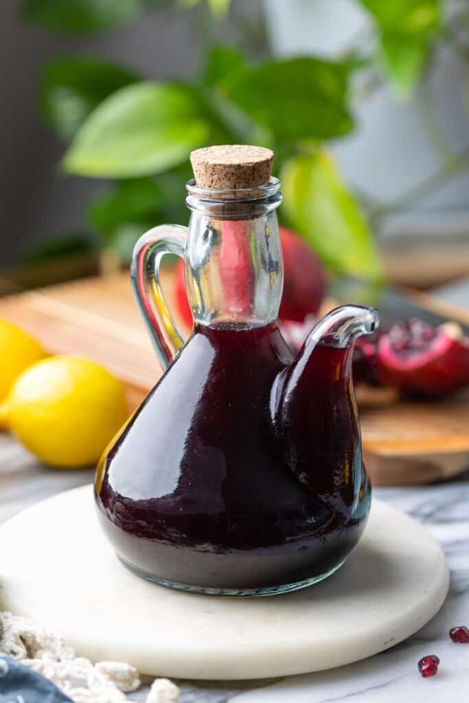 Homemade Grenadine Syrup in a glass drizzling bottle with lemons and pomegranate in the back