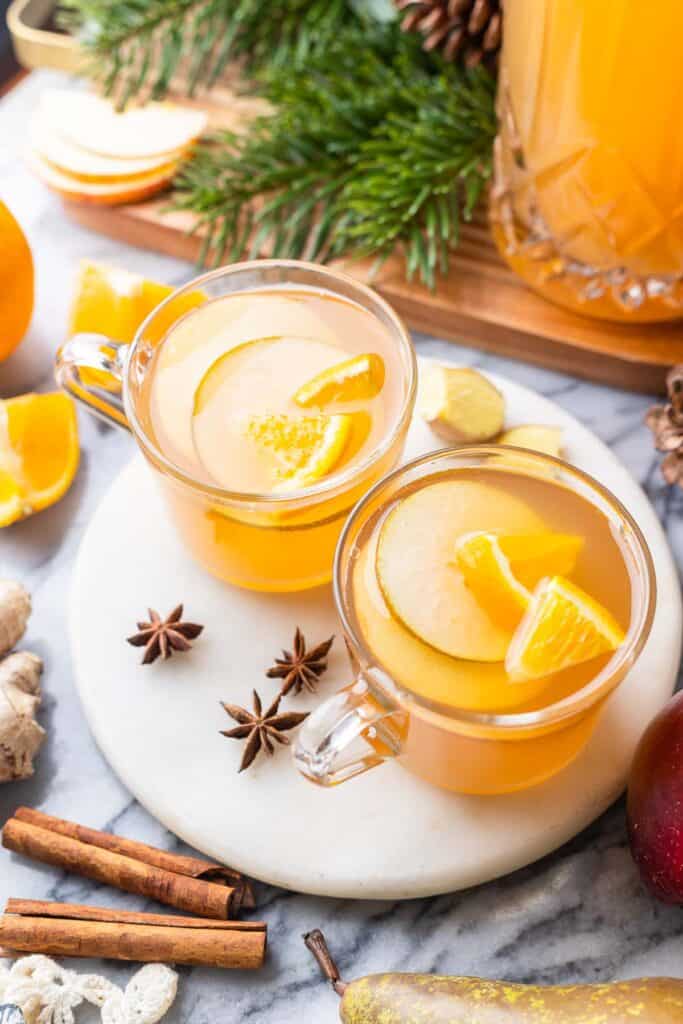 two mugs of apple pear cider on a plate with sliced fruit inside