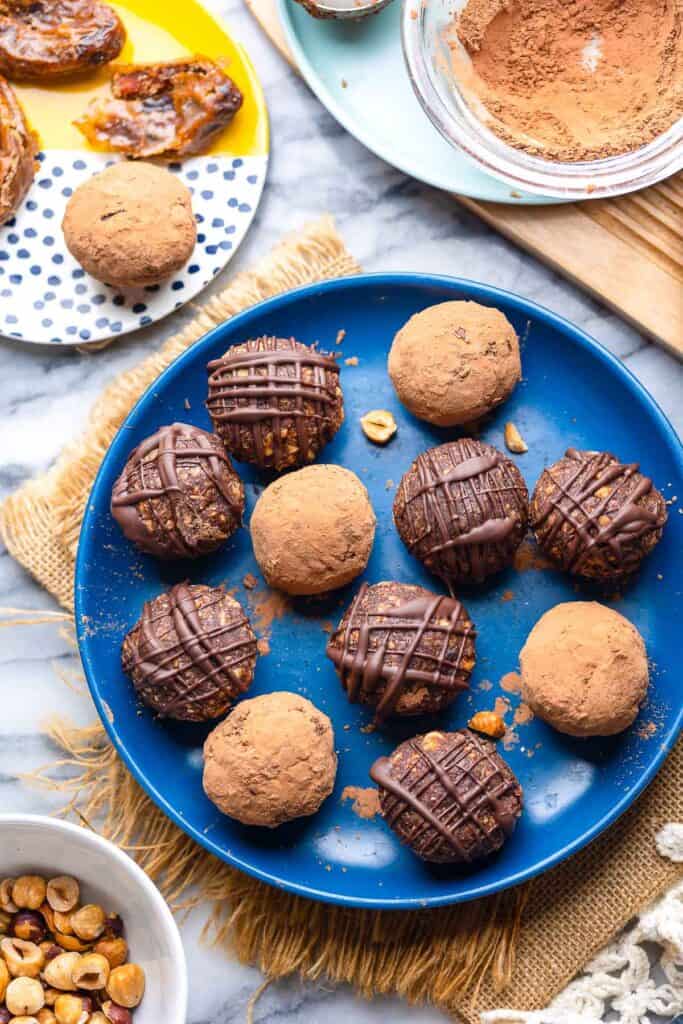 Chocolatey Hazelnut Date Balls covered in cocoa powder and drizzled chocolate on a plate with roasted hazelnuts surounding
