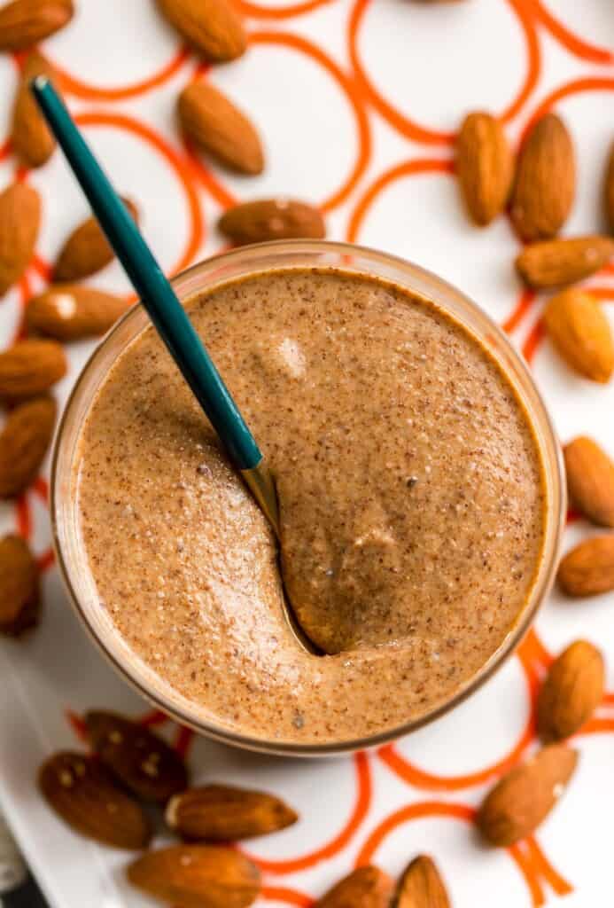 creamy homemade almond butter in a small jar with a spoon on a serving plate surrounded by raw almonds