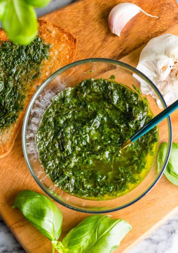 pistou in a small bowl on a serving platter surrounded by garlic, basil leaves and a pieces of toast with pistou spread across