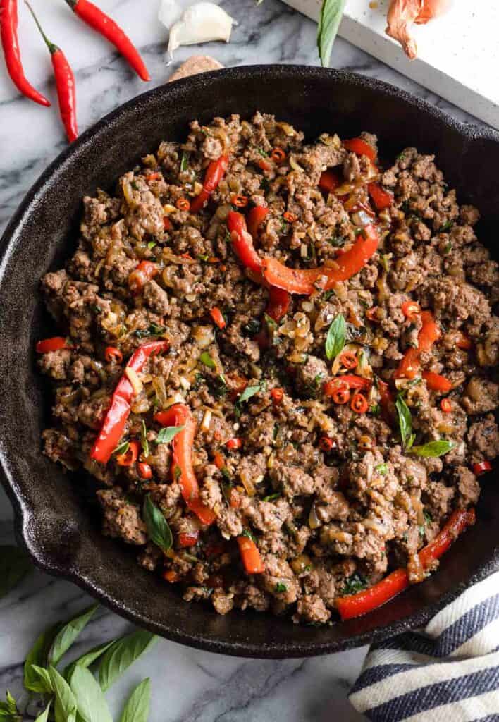 Thai Basil Beef in a cast iron skillet surrounded by the fresh ingredients (thai chilies, basil, garlic)